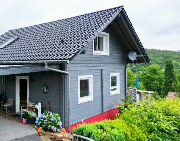 Traditionelles Blockbohlenhaus mit moderner grauer Fassade und roten Akzenten, umgeben von üppigem Grün, präsentiert von FINNHOLZ.