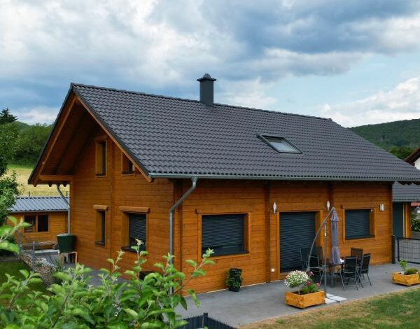 Warmes, honigfarbenes Blockbohlenhaus von FINNHOLZ mit dunklem Dach, vor einem Hintergrund von grünen Hügeln und bewölktem Himmel.