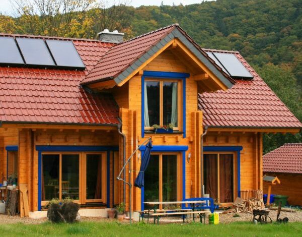 Blockbohlenhaus von FINNHOLZ in leuchtendem Orange mit Solarpaneelen und blauen Akzenten, umgeben von einem naturbelassenen Garten und dicht bewaldeten Hügeln.