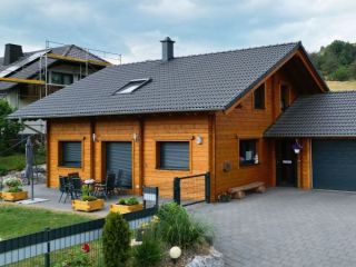 Moderne FINNHOLZ Blockbohlenhaus-Konstruktion mit dunklem Dach, gepflegtem Garten und angeschlossener Garage.