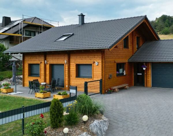 Moderne FINNHOLZ Blockbohlenhaus-Konstruktion mit dunklem Dach, gepflegtem Garten und angeschlossener Garage.