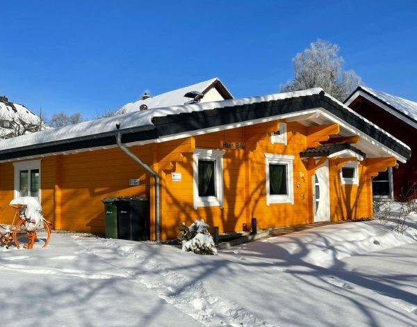 Schneebedecktes FINNHOLZ Blockbohlenhaus mit orangefarbener Fassade und blauem Winterhimmel im Hintergrund.