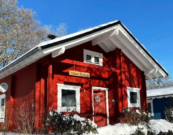 Rotgefärbtes FINNHOLZ Blockbohlenhaus mit dem Namen 'Haus-Ronja' im Winter, umgeben von schneebedeckten Bäumen und klarem blauen Himmel.