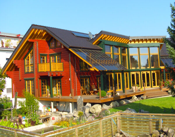 Ein FINNHOLZ Blockhaus mit roter Holzfassade und einem beeindruckenden Glasanbau umgeben von einem wunderschön angelegten Garten.