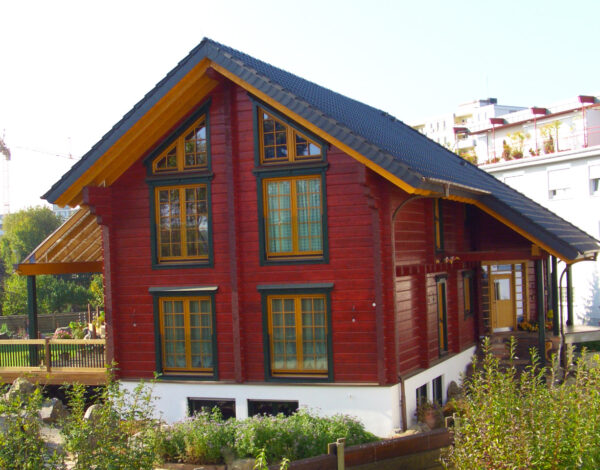 Ein rotes FINNHOLZ Blockhaus mit gelben Fensterrahmen und detaillierter Holzarbeit, eingebettet in ein städtisches Umfeld.