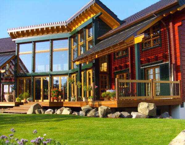 Ein hochwertiges FINNHOLZ Blockbohlenhaus mit charakteristischen roten Wänden, großer Glasfassade und einer einladenden Holzterrasse, eingebettet in eine idyllische Gartenlandschaft.