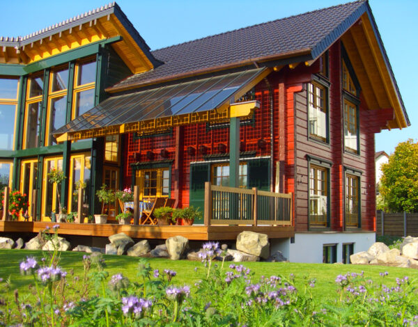 Charmantes FINNHOLZ Blockbohlenhaus mit traditioneller roter Fassade und modernen großen Fenstern, umgeben von einem Garten mit blühenden Pflanzen.