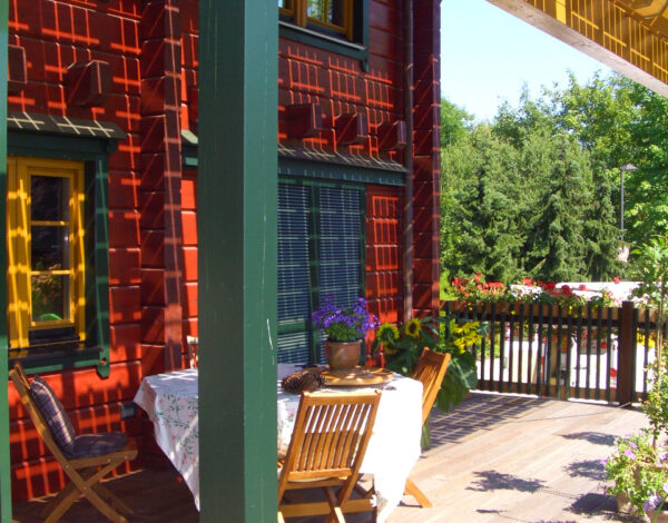 Gemütliche Terrasse eines FINNHOLZ Blockbohlenhauses mit einladendem Esstisch, umgeben von reich blühenden Pflanzen und traditioneller roter Holzfassade.