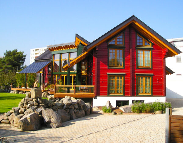 Attraktives rotes FINNHOLZ Blockhaus mit markanten Holzsäulen und einer Steingartenlandschaft vor einem städtischen Hintergrund.