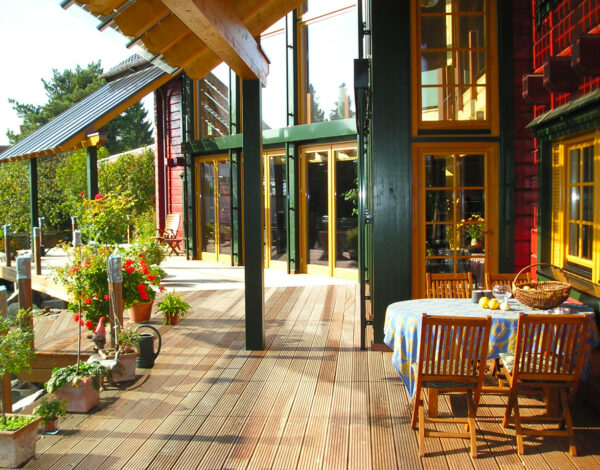 Sonnenbeschienene Terrasse eines FINNHOLZ Blockhauses mit detailreicher roter Holzfassade und einladendem Frühstückstisch.