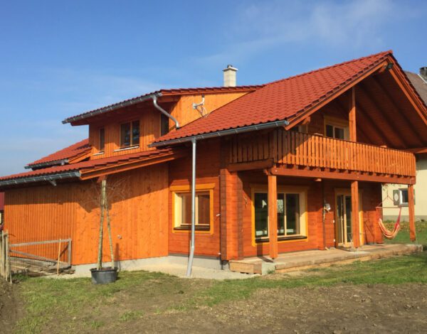 Leuchtendes FINNHOLZ Blockbohlenhaus in Orange mit Hängematte auf der Veranda und klarem Himmel.