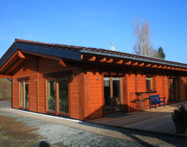 Orange gestrichenes FINNHOLZ Plusenergie-Blockhaus mit Solarpaneelen und blauer Bank vor dem Eingang.