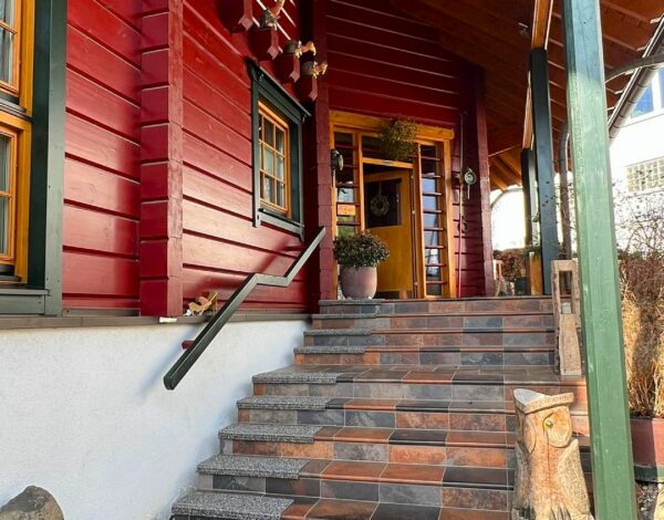 Rot gestrichenes FINNHOLZ Blockbohlenhaus mit einladender Steintreppe und dekorativer Eulenskulptur am Eingang.