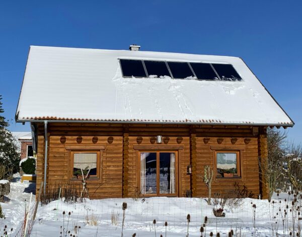 Schneebedecktes Dach eines Rundstammhauses mit Solarmodulen und Blick auf den Garten.