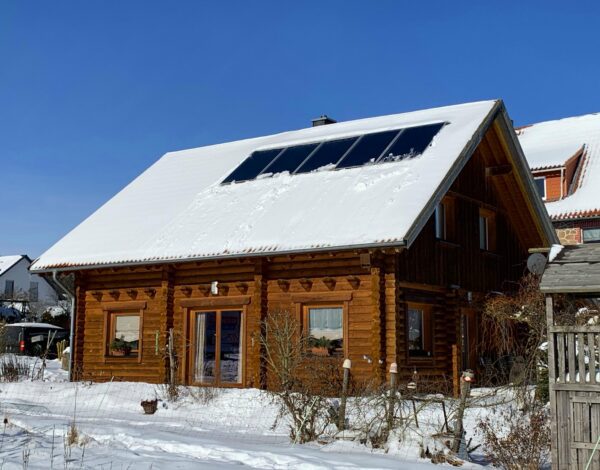 Ansicht eines Blockhauses mit Rundstämmen, Schneebedecktes Dach und Solarmodulen, umgeben von einer winterlichen Landschaft.