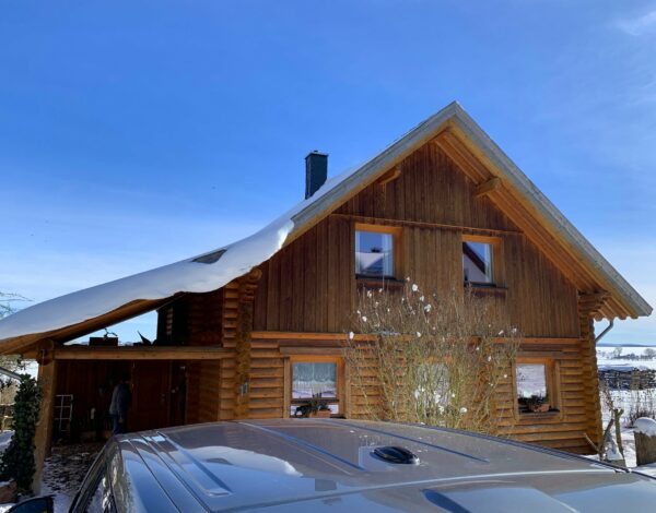 Detailansicht eines Blockhauses mit Rundstämmen und schneebedecktem Dach, Autohaube im Vordergrund.