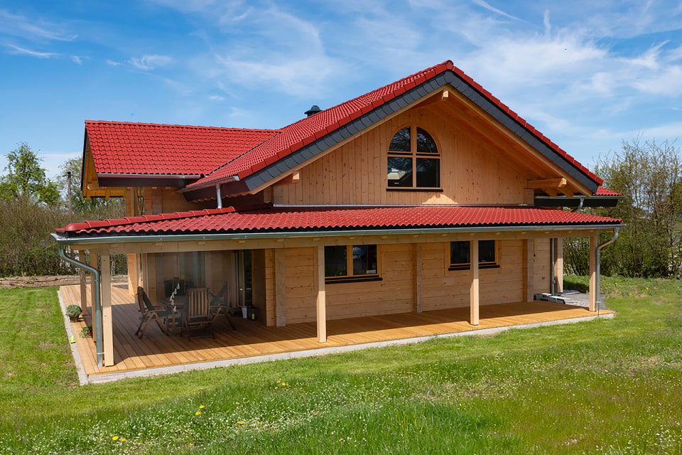 Ein modernes Wohnblockbohlenhaus von FINNHOLZ mit rotem Dach und überdachter Terrasse, umgeben von einer grünen Wiese.