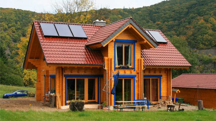 Modernes FINNHOLZ Blockbohlenhaus in Doppelwand-Bauweise mit Solarpaneelen und rotem Dach, umgeben von Natur.