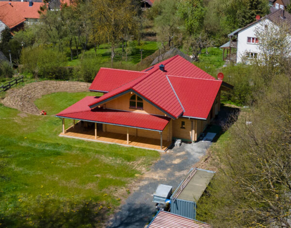 Luftbild eines Blockbohlenhauses mit rotem Dach inmitten einer grünen Wiese repräsentativ für FINNHOLZ Qualität.