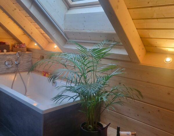 Innenansicht eines FINNHOLZ Blockbohlenhauses mit einer gemütlichen, holzverkleideten Badezimmereinrichtung und einer großen Badewanne, umgeben von natürlichen Holzelementen und einer lebendigen Zimmerpflanze.