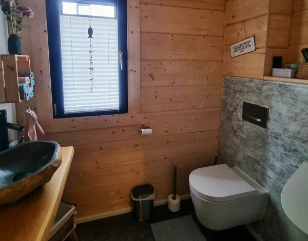 Modernes Badezimmer im FINNHOLZ Blockbohlenhaus mit Natursteinakzenten, rustikalem Holzwaschtisch und natürlicher Holzwandverkleidung, die für eine beruhigende Atmosphäre sorgt.