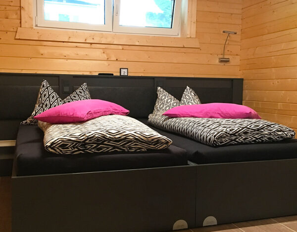 Einladendes Schlafzimmer im FINNHOLZ Blockbohlenhaus, ausgestattet mit zwei Einzelbetten mit schwarzen Bettgestellen und leuchtend pinken Kissen auf einer Wand aus naturbelassenem Holz.
