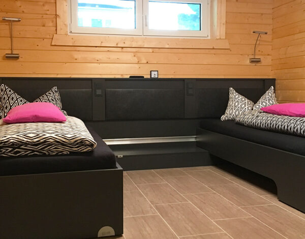 Modern eingerichtetes Schlafzimmer im FINNHOLZ Ferien-Blockbohlenhaus mit zwei eleganten Einzelbetten und dekorativen pinken Kissen auf einer beruhigenden Holzwand.