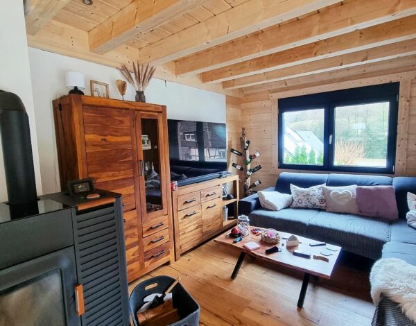 Wohnzimmer in einem FINNHOLZ Blockbohlenhaus mit sichtbaren Holzbalken, einem Kaminofen und stilvollen Möbeln, das eine natürliche und behagliche Wohnatmosphäre ausstrahlt.