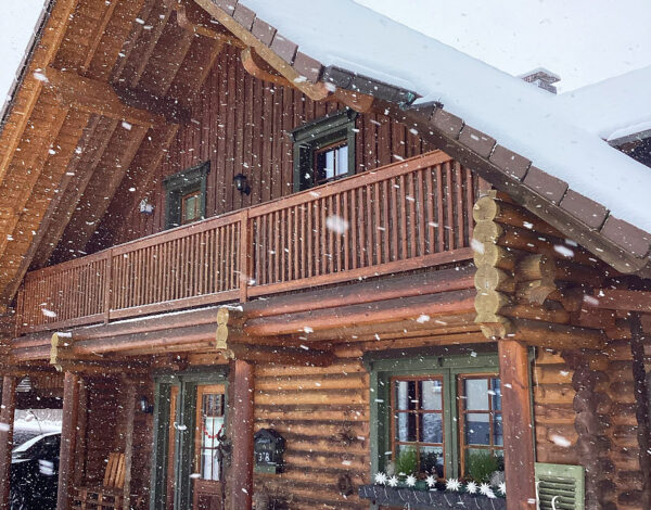 Ein verschneites FINNHOLZ Rundbohlen-Blockhaus in idyllischer Winterlandschaft, mit sichtbaren Holzbohlen und einer einladenden Terrasse.
