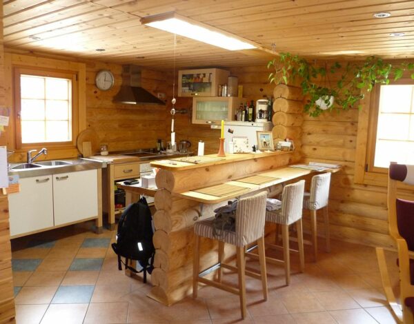 Gemütliche Blockhaus-Küche mit Rundbohlenwänden von FINNHOLZ, ausgestattet mit modernen Küchengeräten und einer langen Holztischgarnitur.