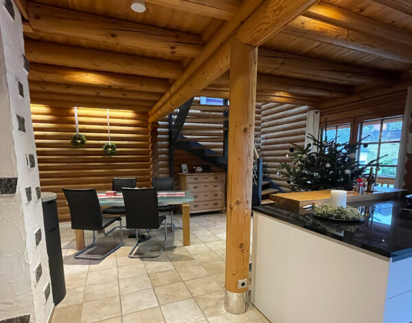 Stilvoller Essbereich im FINNHOLZ Rundbohlen-Blockhaus mit sichtbaren Holzbalken, moderner Küche und einer eleganten Holztreppe.