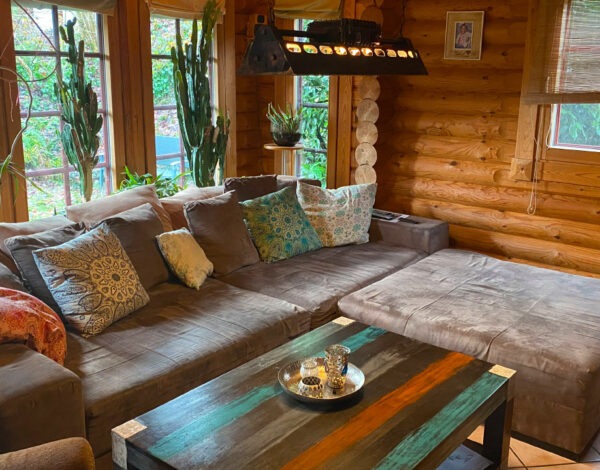 Einladendes Wohnzimmer in einem FINNHOLZ Rundbohlen-Blockhaus, ausgestattet mit einer großzügigen Eckcouch, bunten Kissen und einem handgefertigten Holztisch.