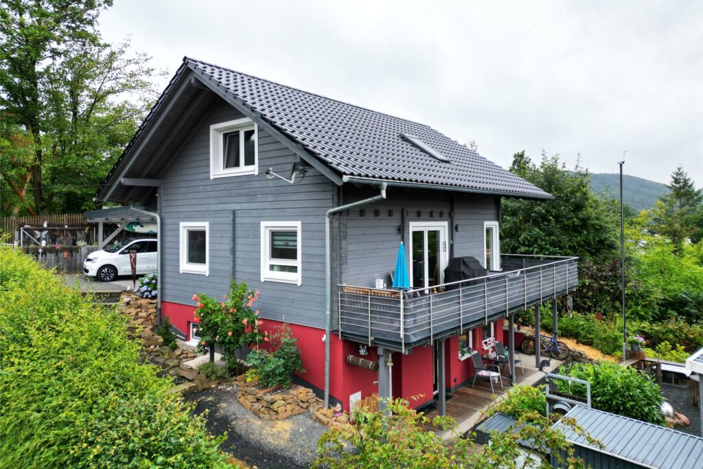 FINNHOLZ Ferienhaus mit roter gestrichenen, verputzten Fassade am unteren Stockwerk und grauen Holzstämmen oben, eingebettet in grüner Natur.