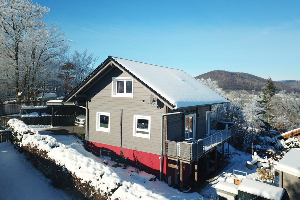 FINNHOLZ Ferienhaus im Winter, umgeben von einer verschneiten Landschaft und mit einer weit entfernten, waldbedeckten Hügel im Hintergrund.