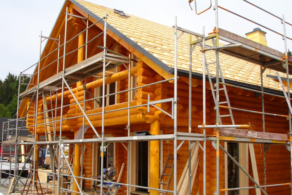 Blockhaus, eingehüllt in Baugerüste, mit Wänden aus mit schützendem Anstrich behandelten Rundstämmen, der dem Holz einen warmen rötlich-orangen Farbton verleiht.