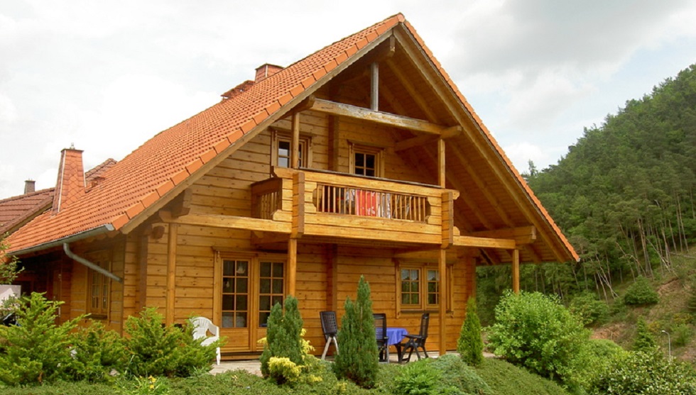 Graues Blockhaus aus Holz mit grünem Dach, umgeben von einer grünen Rasenfläche und Bäumen, unter einem blauem Himmel.