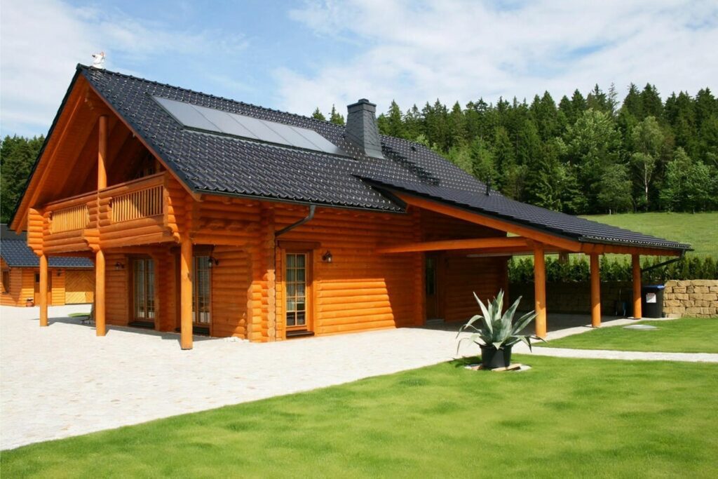Goldenes Finnholz Blockhaus mit Terrasse, umgeben von grünem Rasen und Steinlandschaft, schwarzes Dach, Wald im Hintergrund.