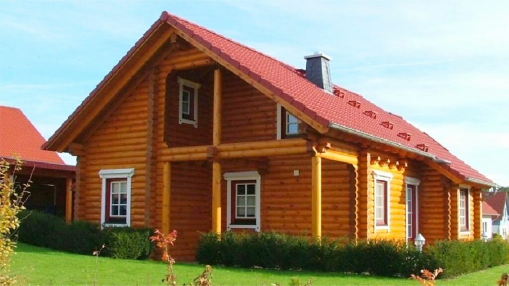 Golden-holzfarbenes Finnholz Blockhaus mit roter Dachschindel, weiß umrandete Fenster und gepflegter grüner Rasen.