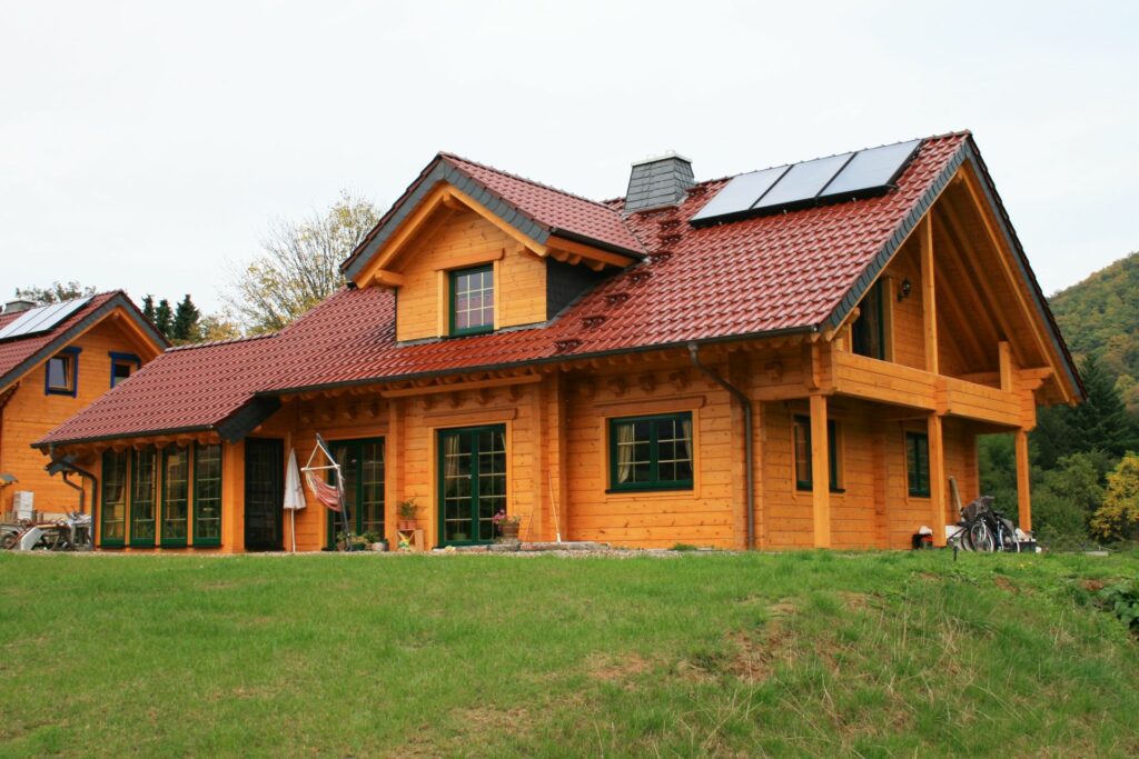 Holzhaus FINNHOLZ mit rotem Dach und Solarpaneelen in grüner Umgebung.
