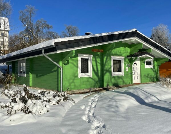 Grünes Blockhaus von Finnholz, aus Kantholz gefertigt, verschneit mit strahlend blauem Himmel im Hintergrund.
