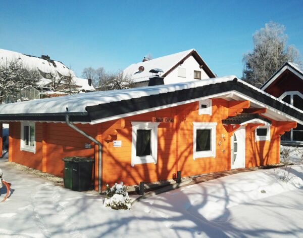 Blockhaus in Orange, bedeckt mit Schnee, im Hintergrund weitere verschneite Häuser.