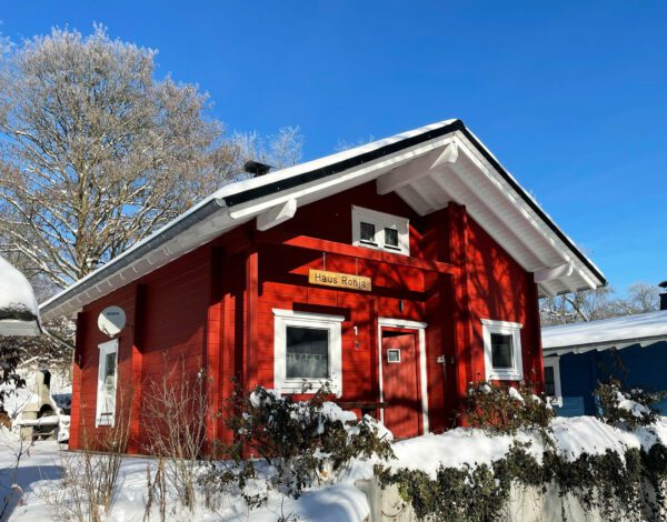 Rotes Blockhaus von Finnholz im Winter, umgeben von verschneiten Sträuchern und Bäumen.
