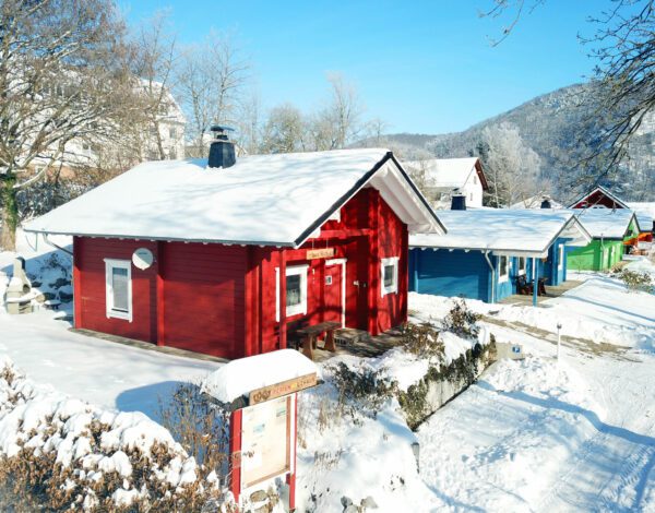 Panoramablick auf ein rotes Finnholz-Blockhaus im Winter, umgeben von Schnee und anderen bunten Häusern.