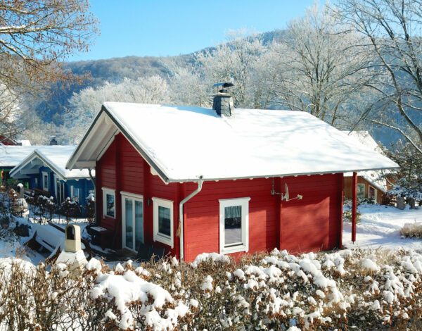 Vorderansicht des roten Finnholz Blockhauses mit verschneitem Dach und winterlicher Umgebung.