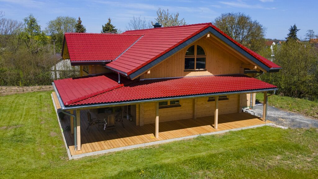 Modernes Blockbohlenhaus mit rotem Dach und überdachter Terrasse auf grüner Wiese.